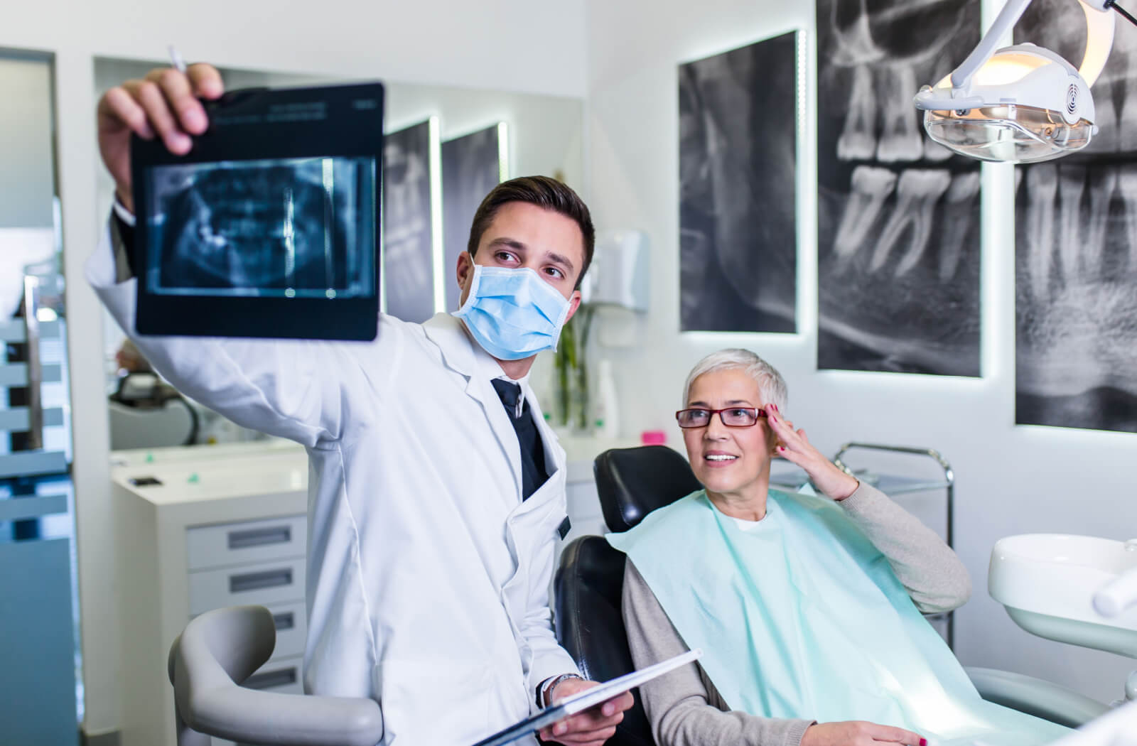 A male dentist with a mask explaining a dental procedure to a senior woman while holding a dental X-ray image