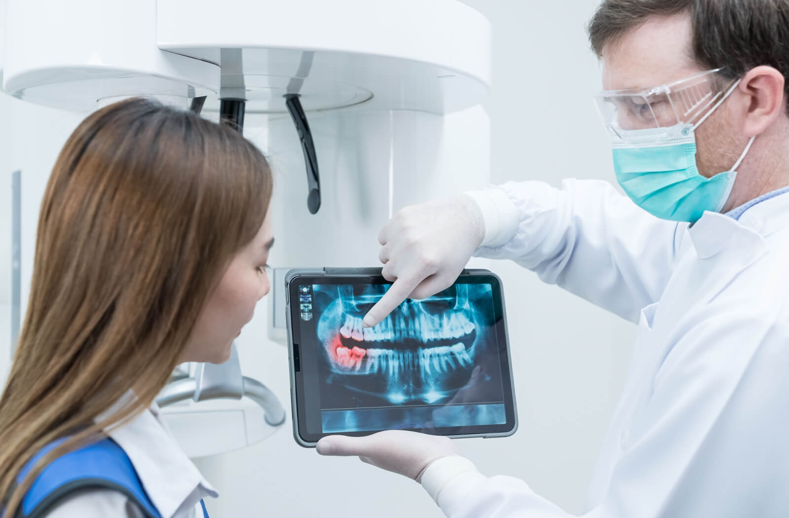 A male dentist explaining a dental procedure to a woman while holding a dental X-ray image.
